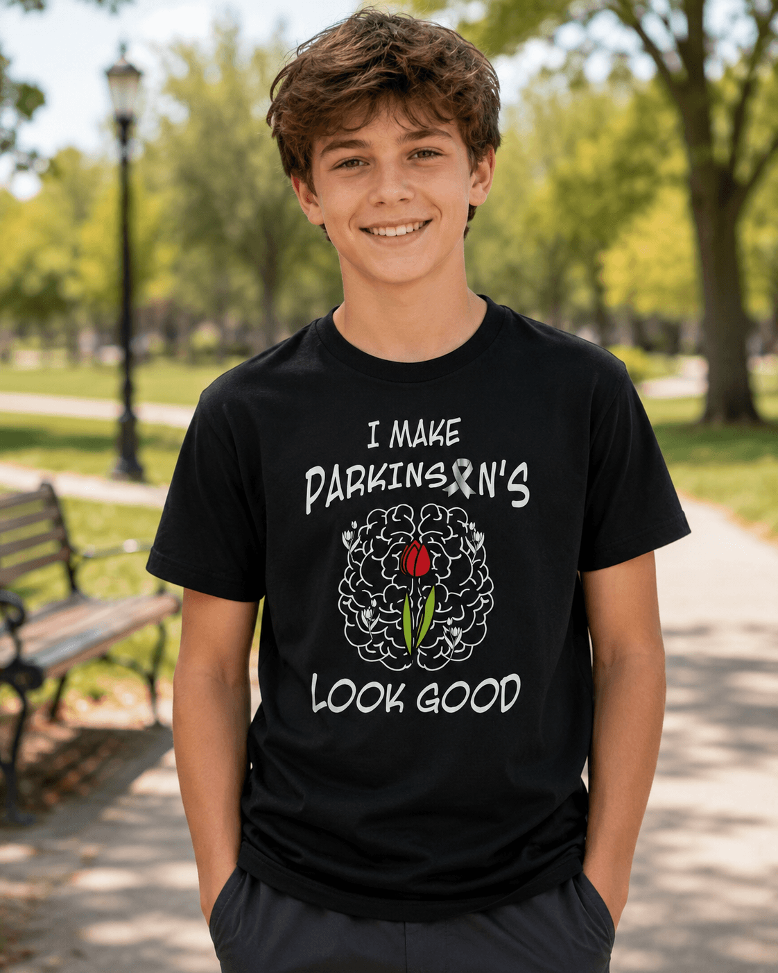 A smiling young man stands outdoors on a park path wearing a black graphic T-shirt. The shirt has white text reading 'I MAKE PARKINSON'S LOOK GOOD' and a brain illustration with a red rose graphic in the center.