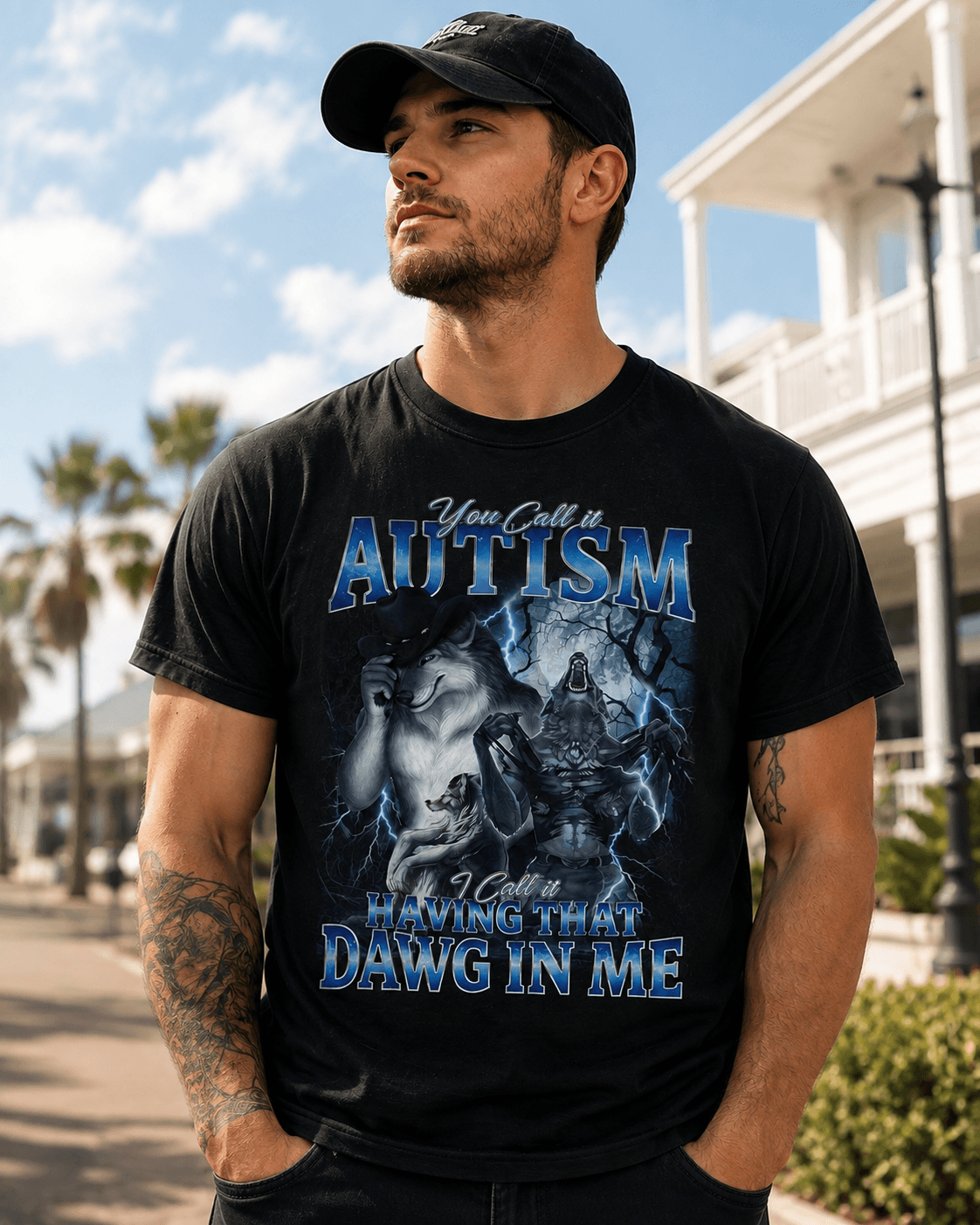 A man wearing a black graphic t-shirt with a blue and white design that reads 'You Call It Autism I Call It Having That Dawg In Me', standing outdoors on a sunny day with a beachfront building in the background.
